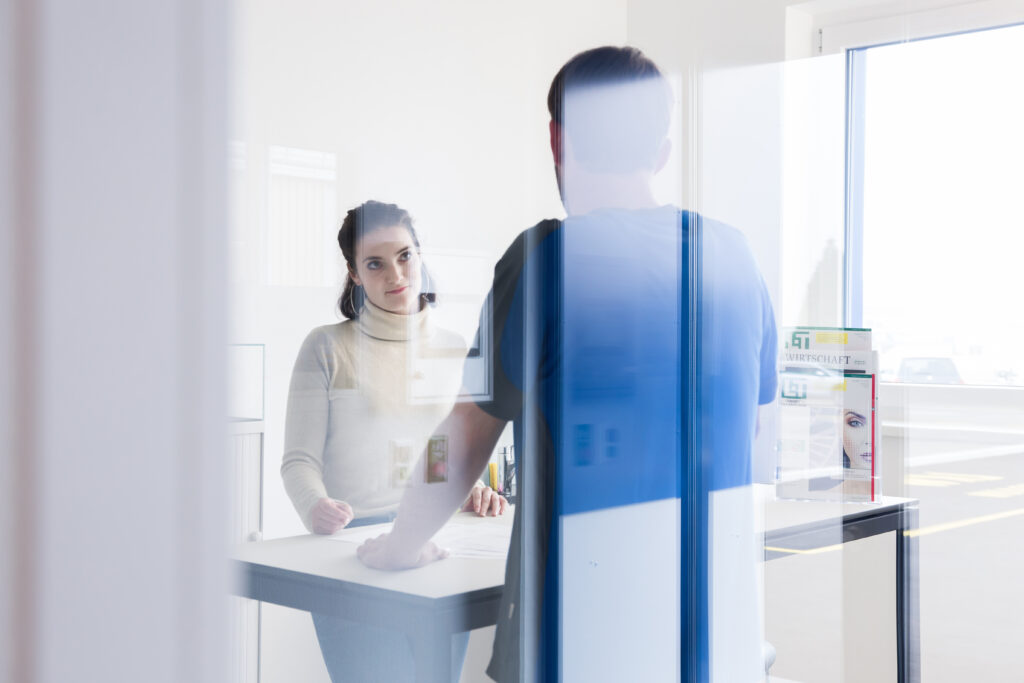 Zwei Fachpersonen von Primcut im Besprechungsraum, durch eine Glasscheibe mit Spiegelung. Sie besprechen einen Druckauftrag. Das Bild ist Weiss und Blau getönt.
